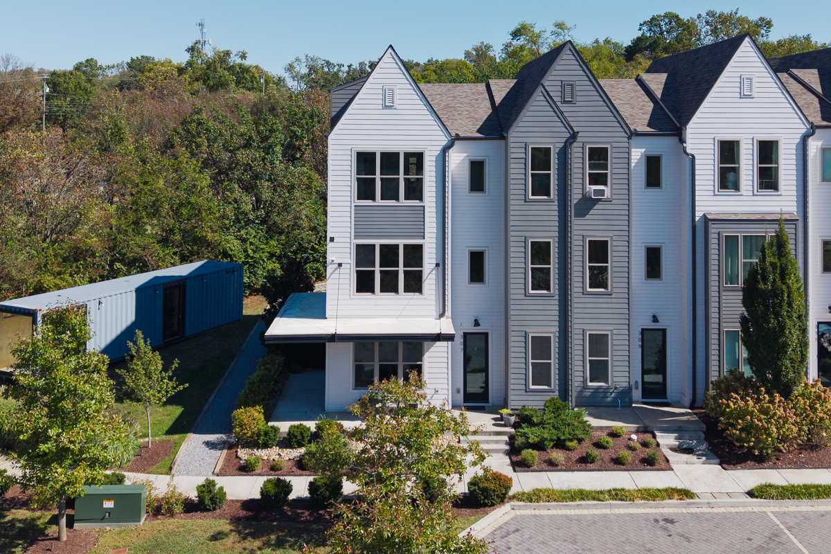 A row of modern townhomes at 307 E Village Ln with white and gray siding, featuring a paved parking area and a blue shipping container nearby.