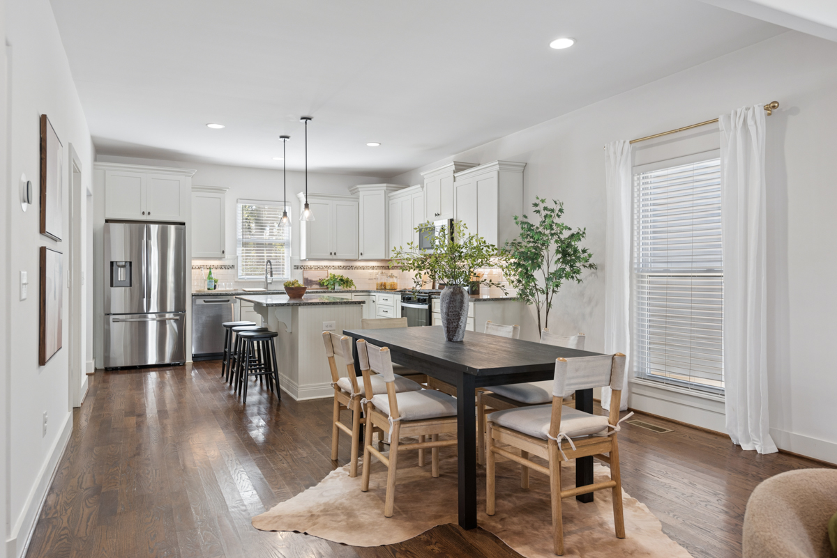 A dining room at 6363 Columbia Ave with a dark wooden table and four chairs atop a rug, adjacent to an open kitchen with white cabinets and stainless steel appliances.