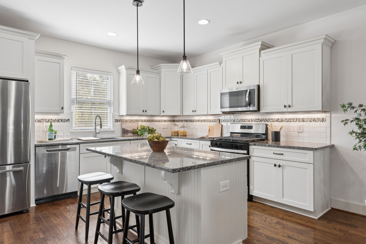 A white kitchen in this Nashville real estate listing featuring a center island, granite countertops, stainless steel appliances, and three black bar stools.