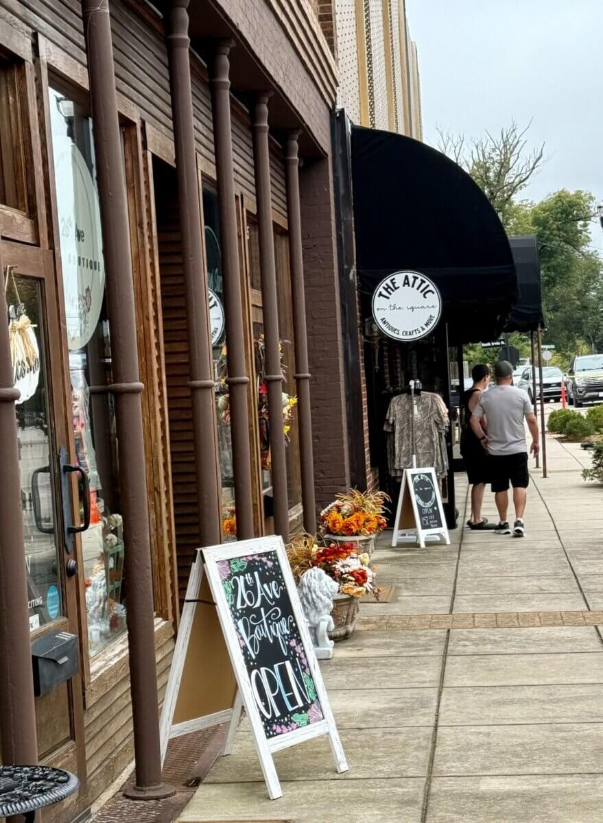 Two people walk past shops in a historic downtown, passing a sidewalk sign that reads 26th Ave Boutique Open.