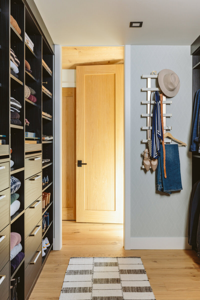 A walk-in closet with floor-to-ceiling dark wood shelving, a central door, and a rack displaying winter clothes, demonstrating effective closet organization with a hat, scarf, heels, and jeans.