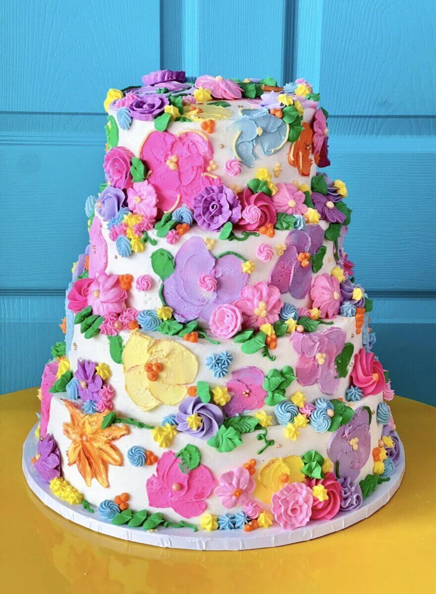 Four-tier wedding cake covered in colorful buttercream flowers against a bright blue door