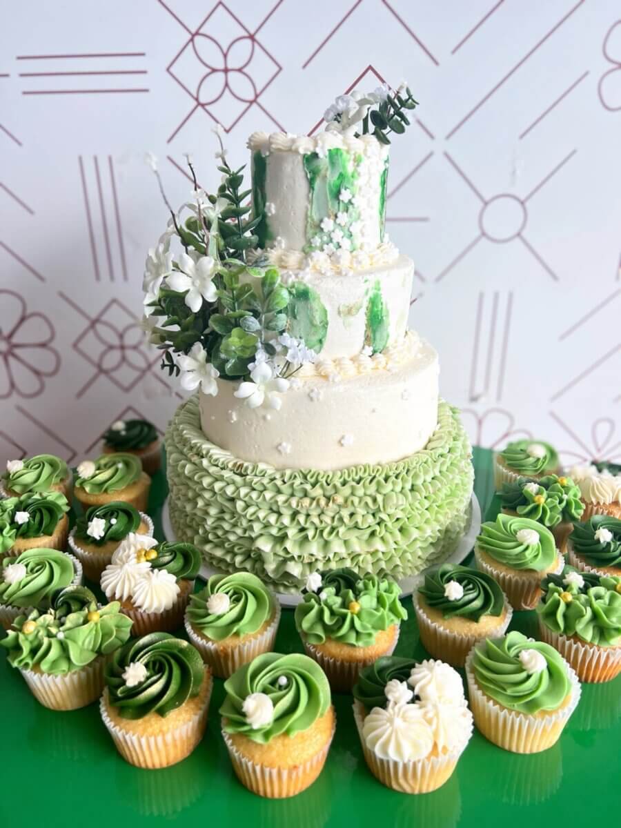 Three-tier white wedding cake with green watercolor accents, a ruffled base and faux greenery, surrounded by green cupcakes