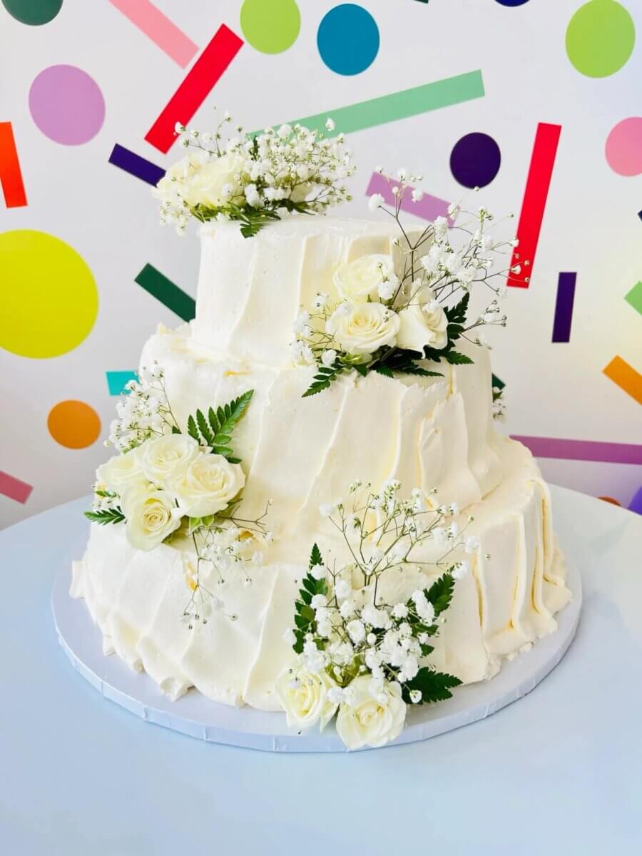 Three-tier white buttercream wedding cake with fresh white roses and baby's breath against a colorful confetti wall