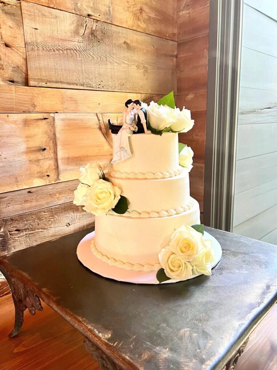 Three-tier white wedding cake with fresh white roses and a bride and groom topper on a rustic wood background